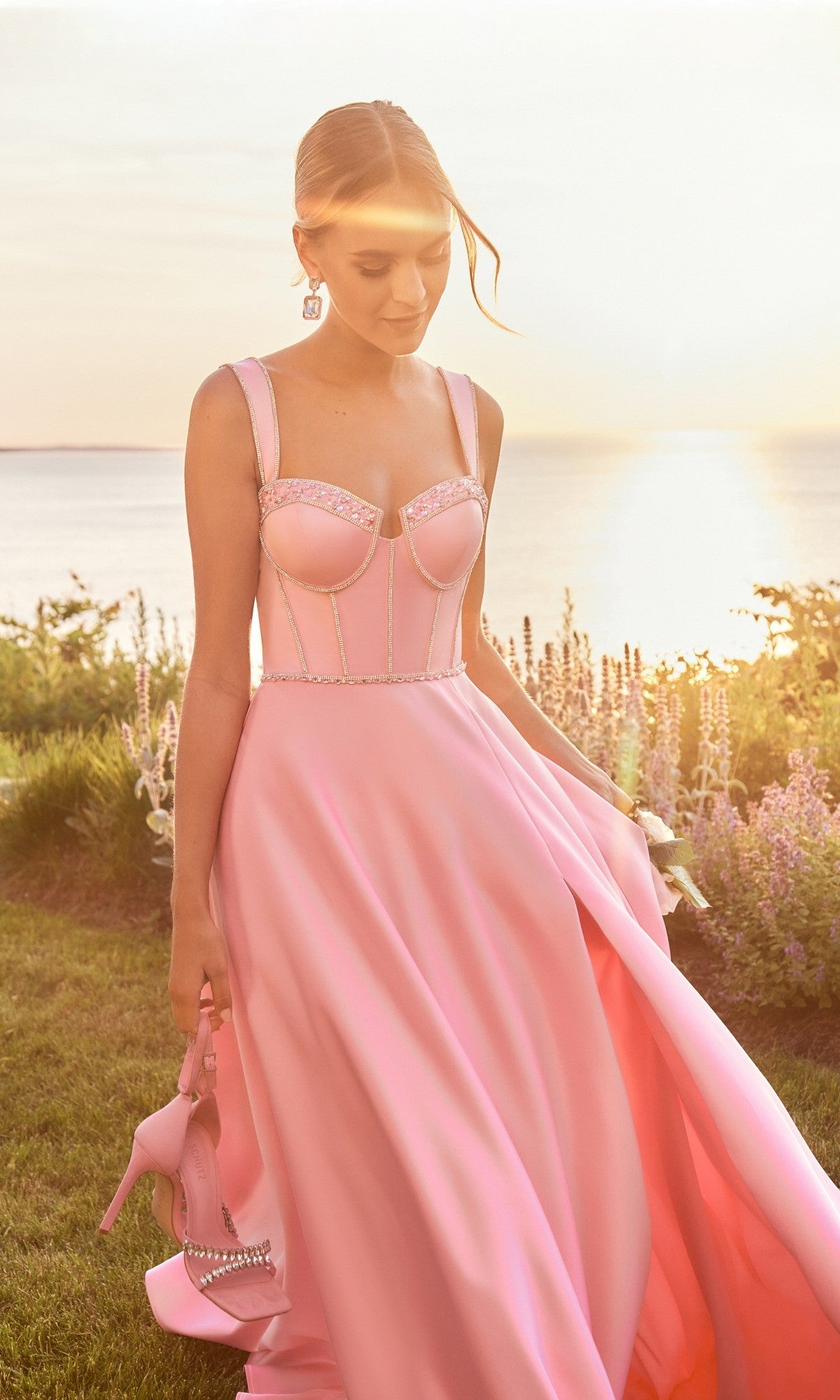 A woman in the Morilee 49177 long prom dress holds matching heels while standing on grass near water at sunset, surrounded by flowers and greenery.